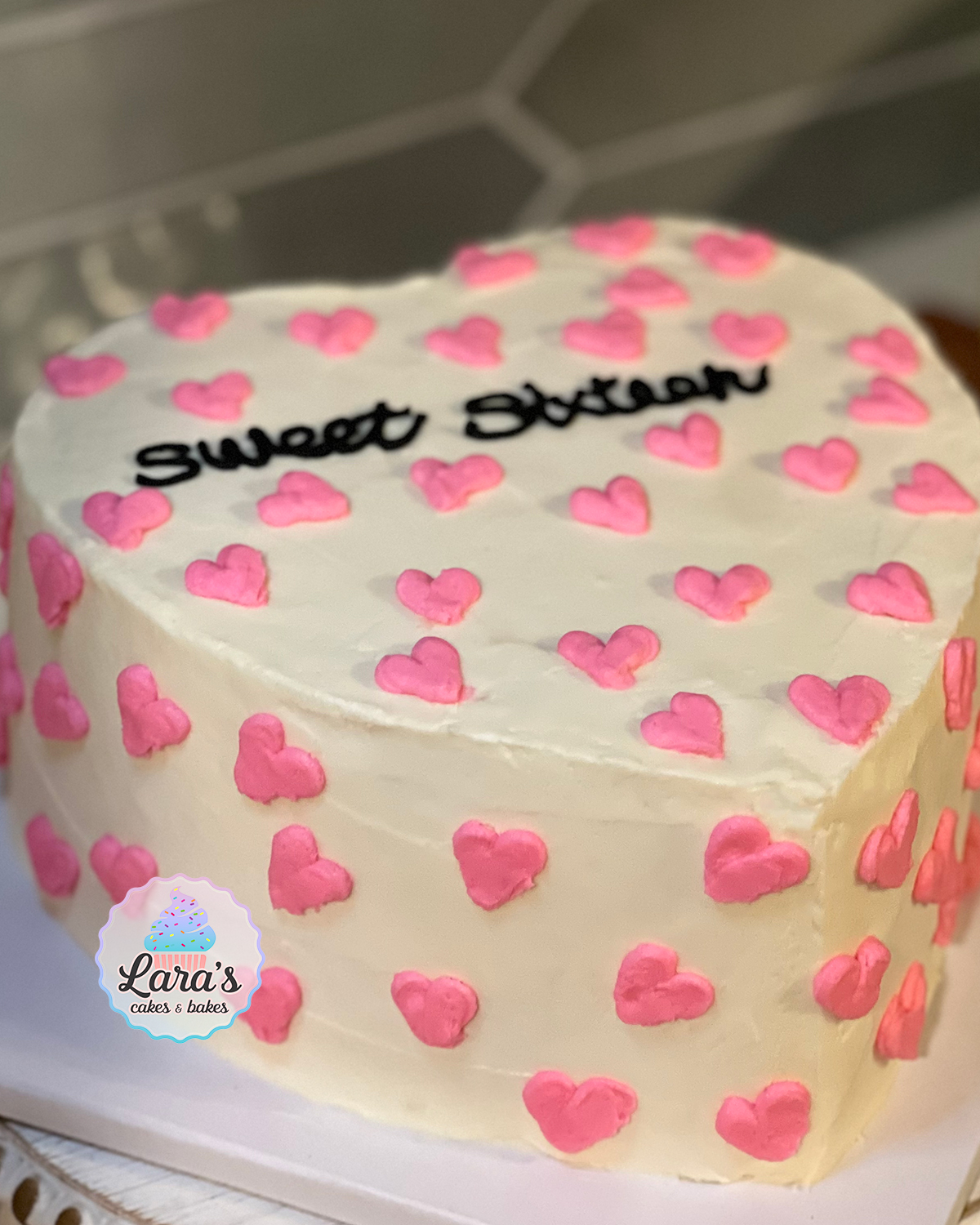 Heart shaped cake with pink hearts covering cake and "sweet sixteen" writing in black
